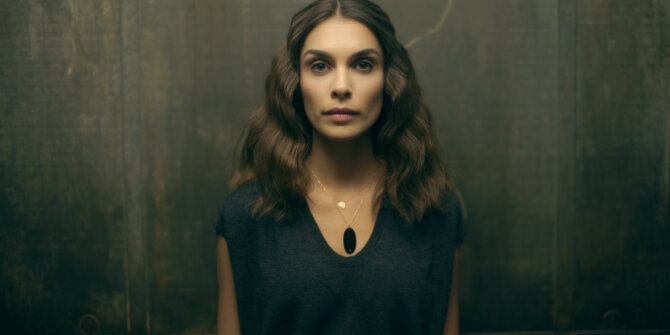 Woman with wavy brown hair wearing a black pendant against a dark wall – Ginette NY Chiaroscuro